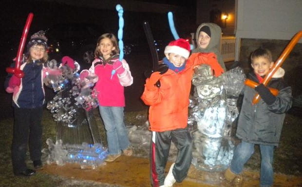 Children in winter gear posing around a lit holiday ice sculpture display