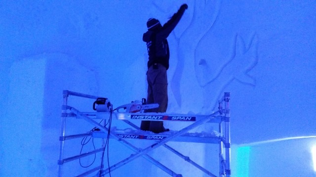 Artist on scaffolding refining an ice structure under vibrant blue event lighting