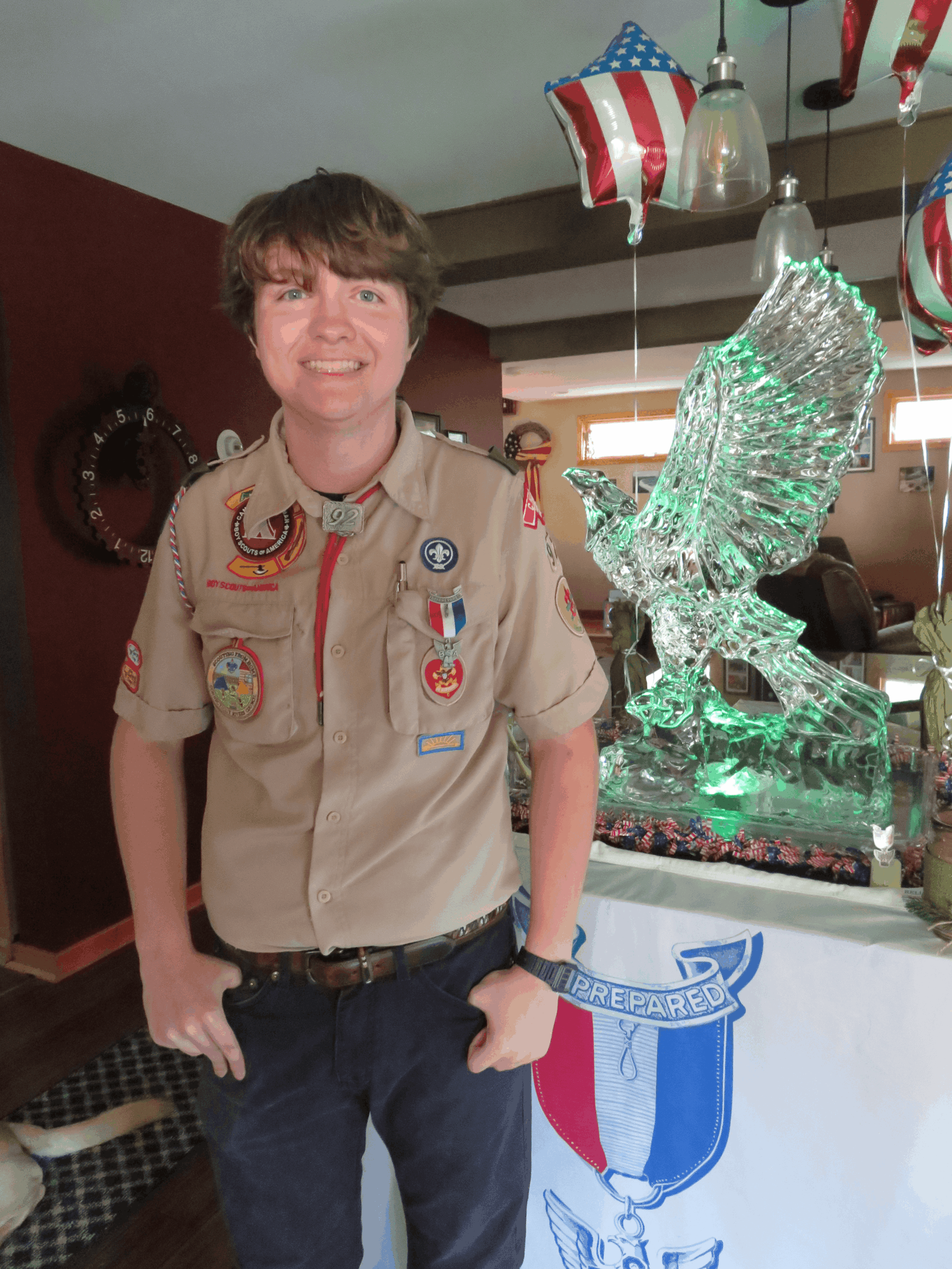 Boy scout standing indoors next to a large detailed green sculpture installation
