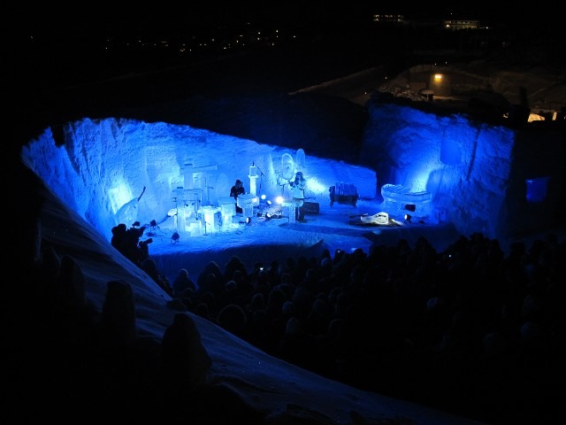 Wide shot of a winter ice festival village glowing at night under deep blue lighting