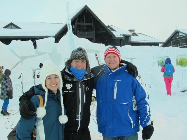 Three smiling people in winter clothing posing in front of a snowy building