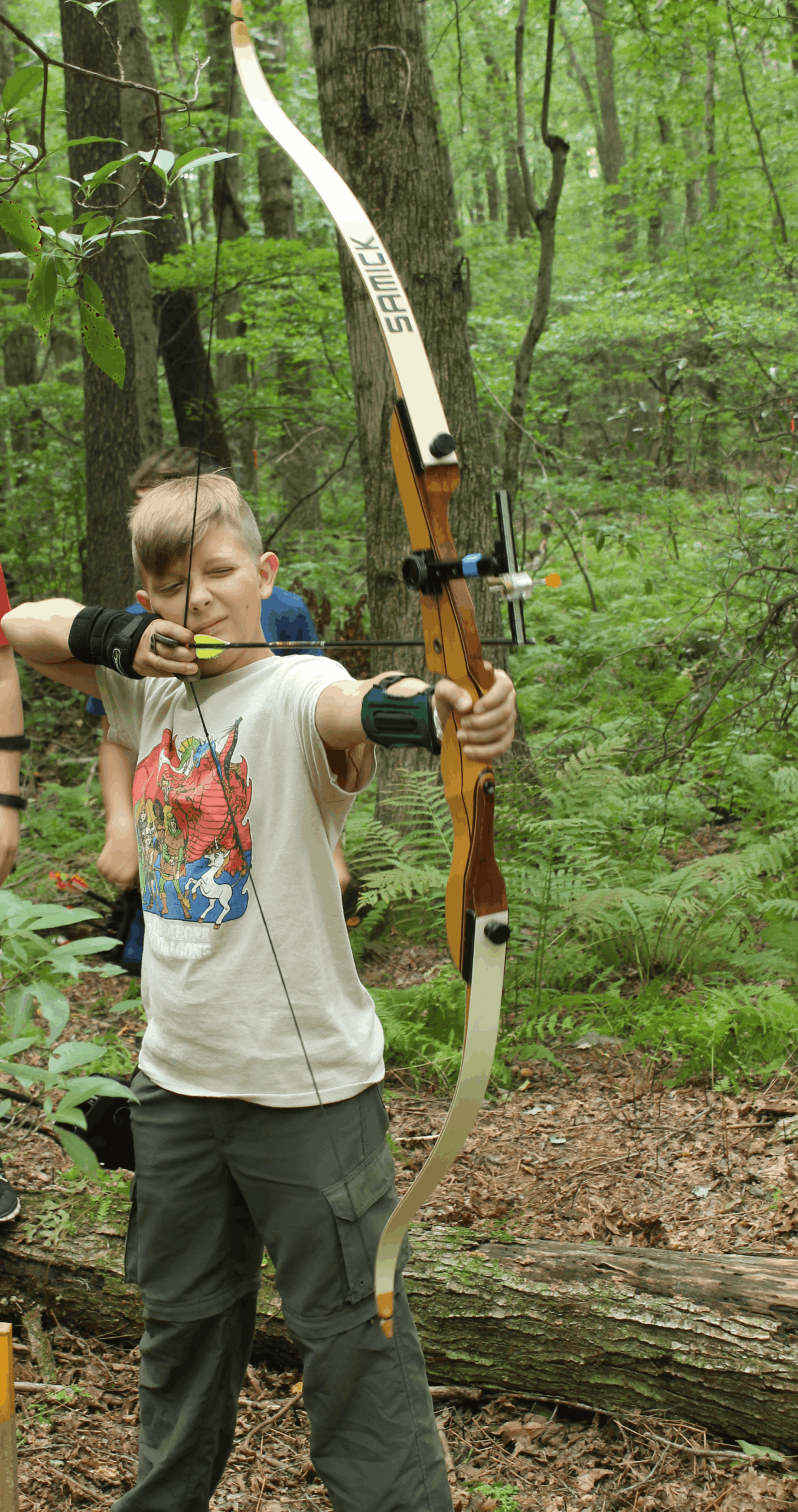 Young boy aiming a bow and arrow outdoors in a lush green forest